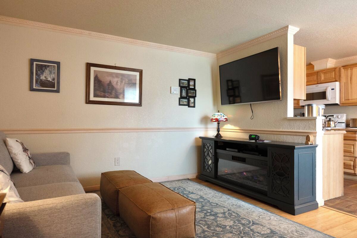 A living room with a sofa, ottoman, and a fireplace. There is a kitchen area with wooden cabinets and a microwave.