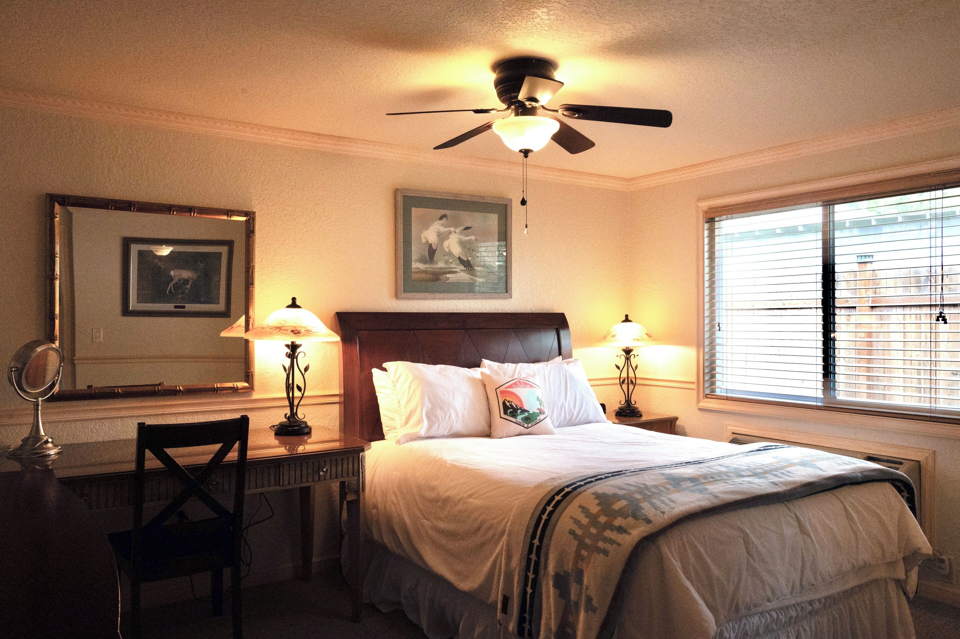A bedroom with a bed, two bedside lamps, a ceiling fan, a window with blinds, a chair, a desk with a mirror, and framed pictures on the wall.