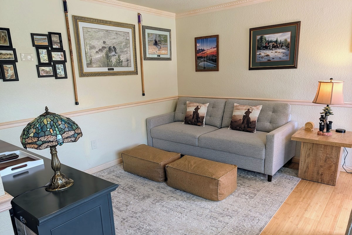 A living room with a grey sofa, two ottomans, a lamp, a small table, and framed pictures on the wall.