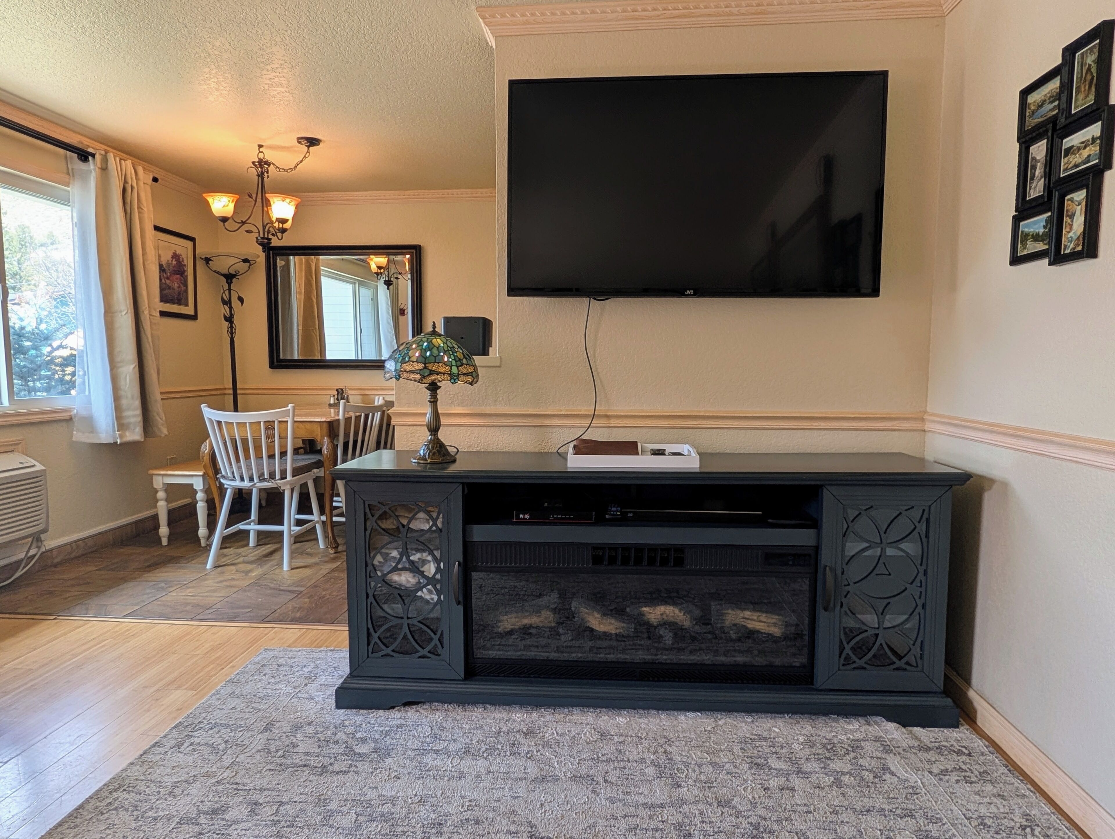 A living room with a flat-screen TV mounted on the wall, a fireplace, a dining table with chairs, and a chandelier.