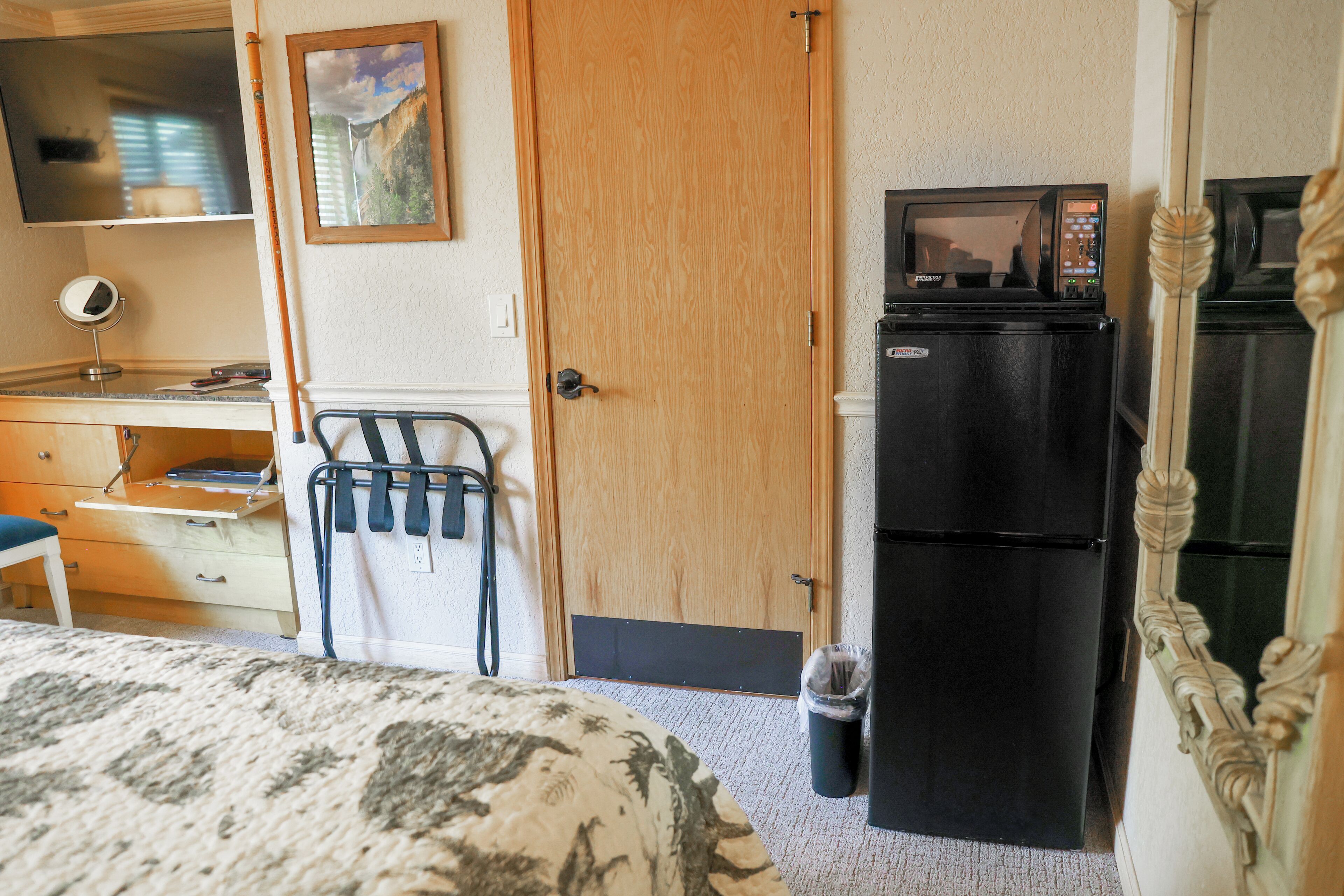 A hotel room with a bed, a desk with a computer, a refrigerator, a microwave, a trash bin, and a mirror.