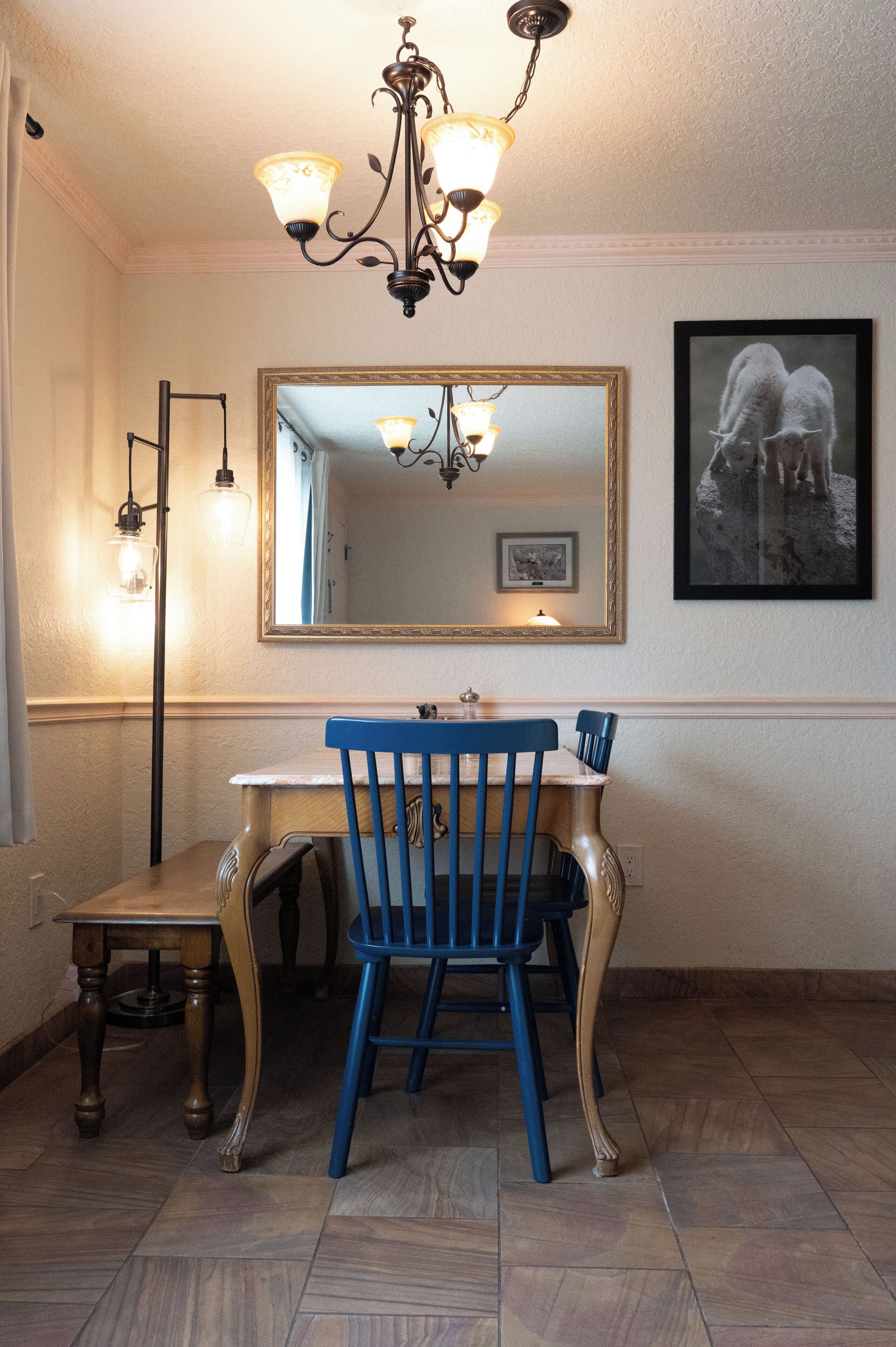 A dining area with a wooden table, a blue chair, a chandelier, and a framed mirror on the wall.
