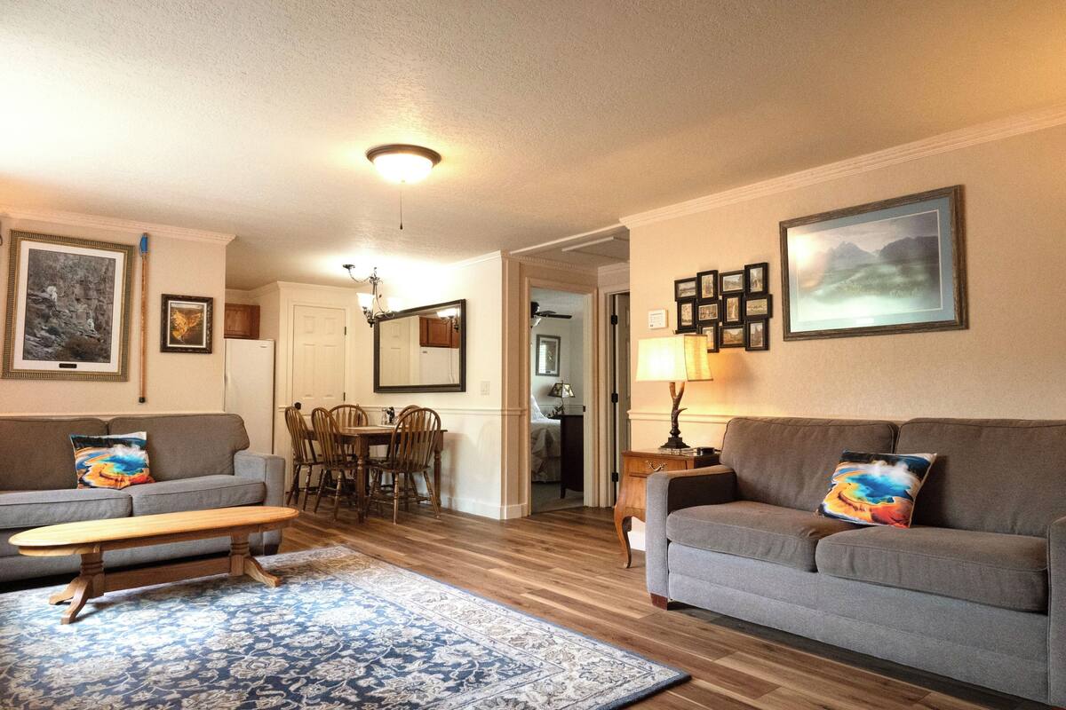 A living room with a sofa, a coffee table, a dining table with chairs, and a kitchen area in the background.
