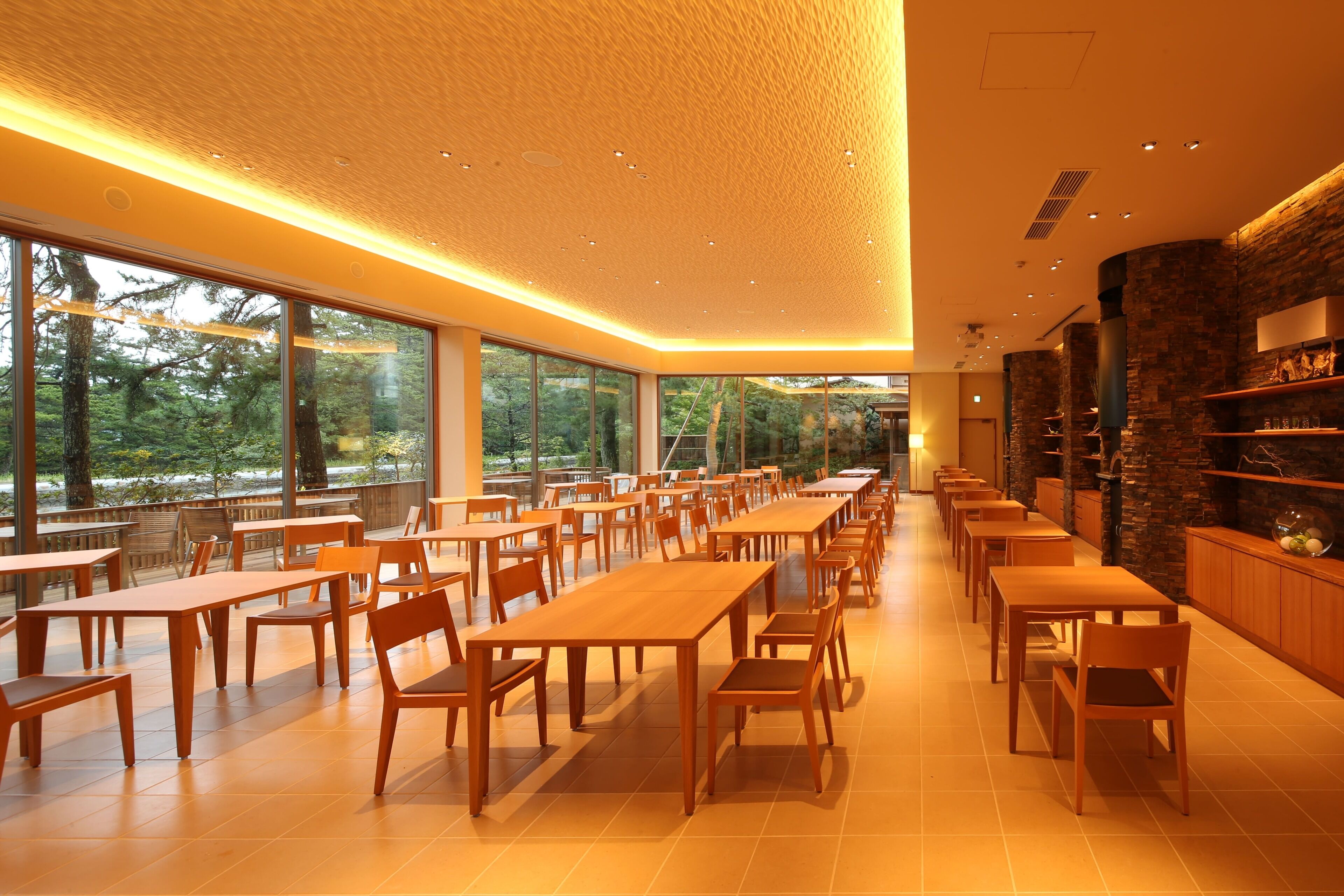 A spacious dining area with long wooden tables and chairs, large windows offering a view of trees, and a warm ceiling with recessed lighting.