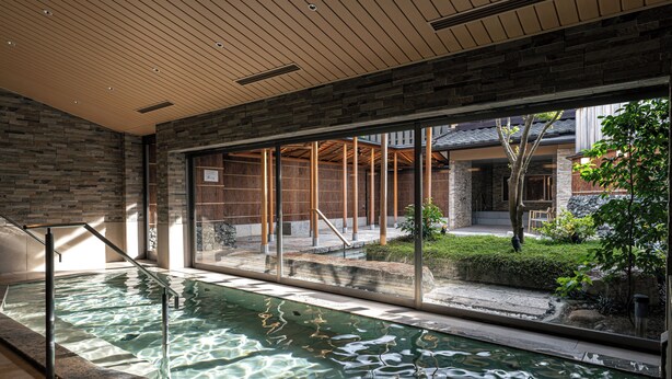 An indoor swimming pool with clear water, wooden ceiling, and large windows offering a view of an outdoor garden and building. - Monjusou (Miyazu)