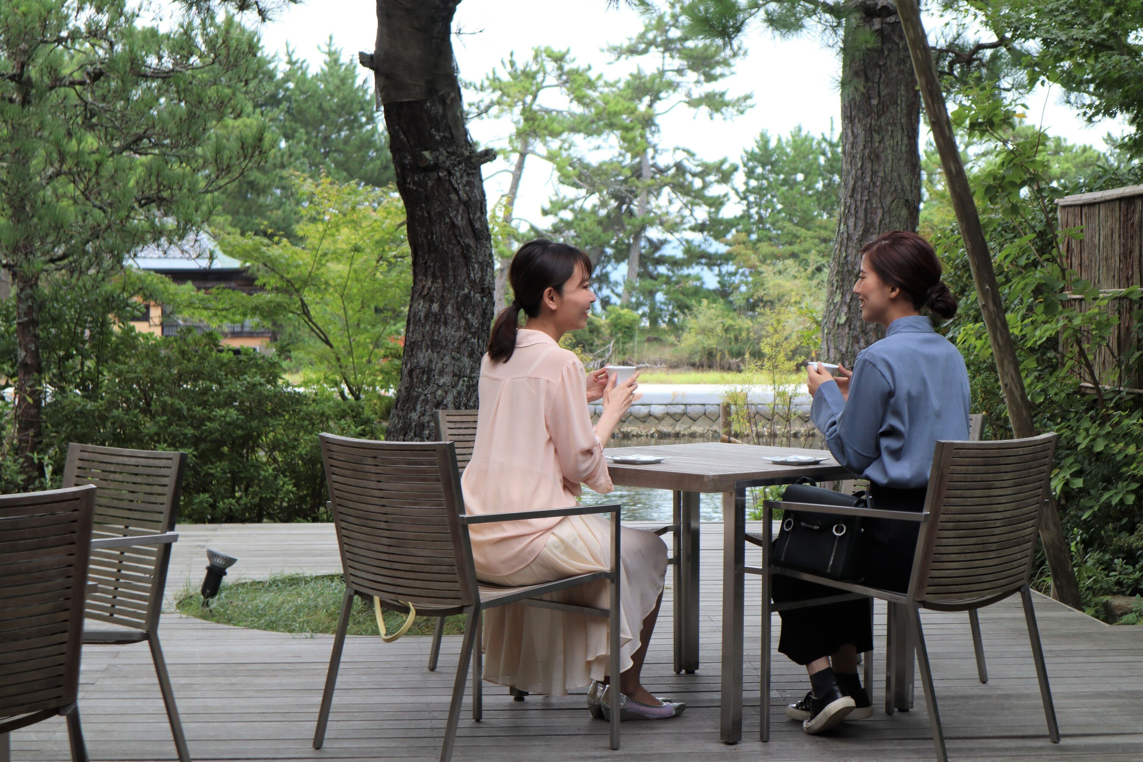 Two people seated at a table outdoors with chairs and a table, surrounded by trees and a fence.