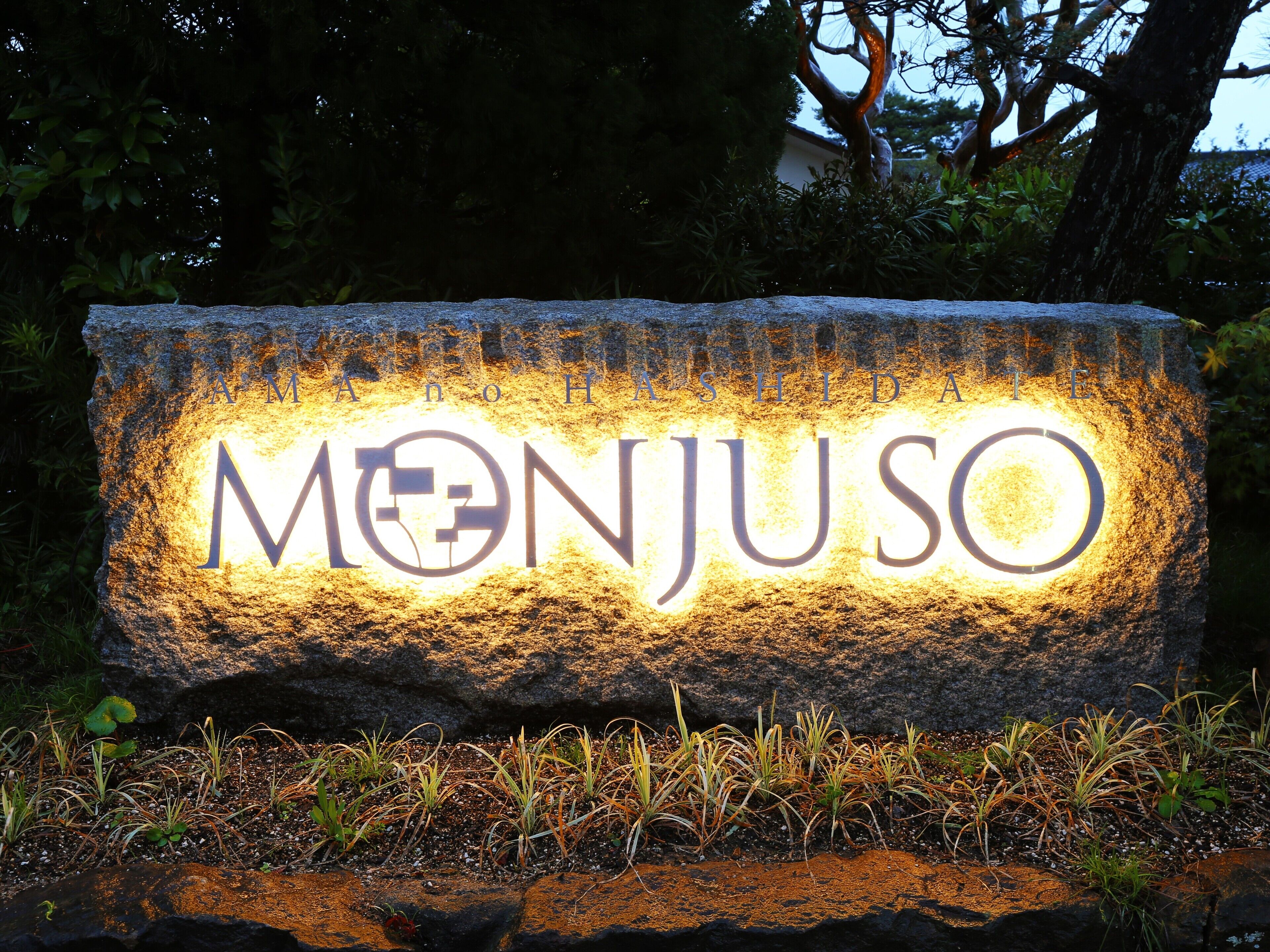 A stone sign with the name "MONJUSO" illuminated in the evening.