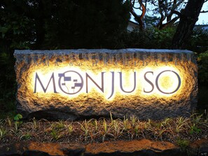 A stone sign with the name "MONJUSO" illuminated in the evening.