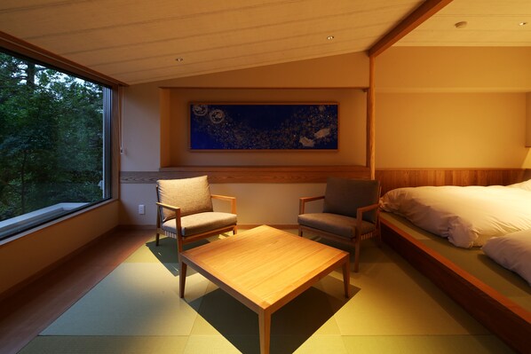 A modern hotel room with a large window, a bed, two chairs, a small table, and a painting on the wall.