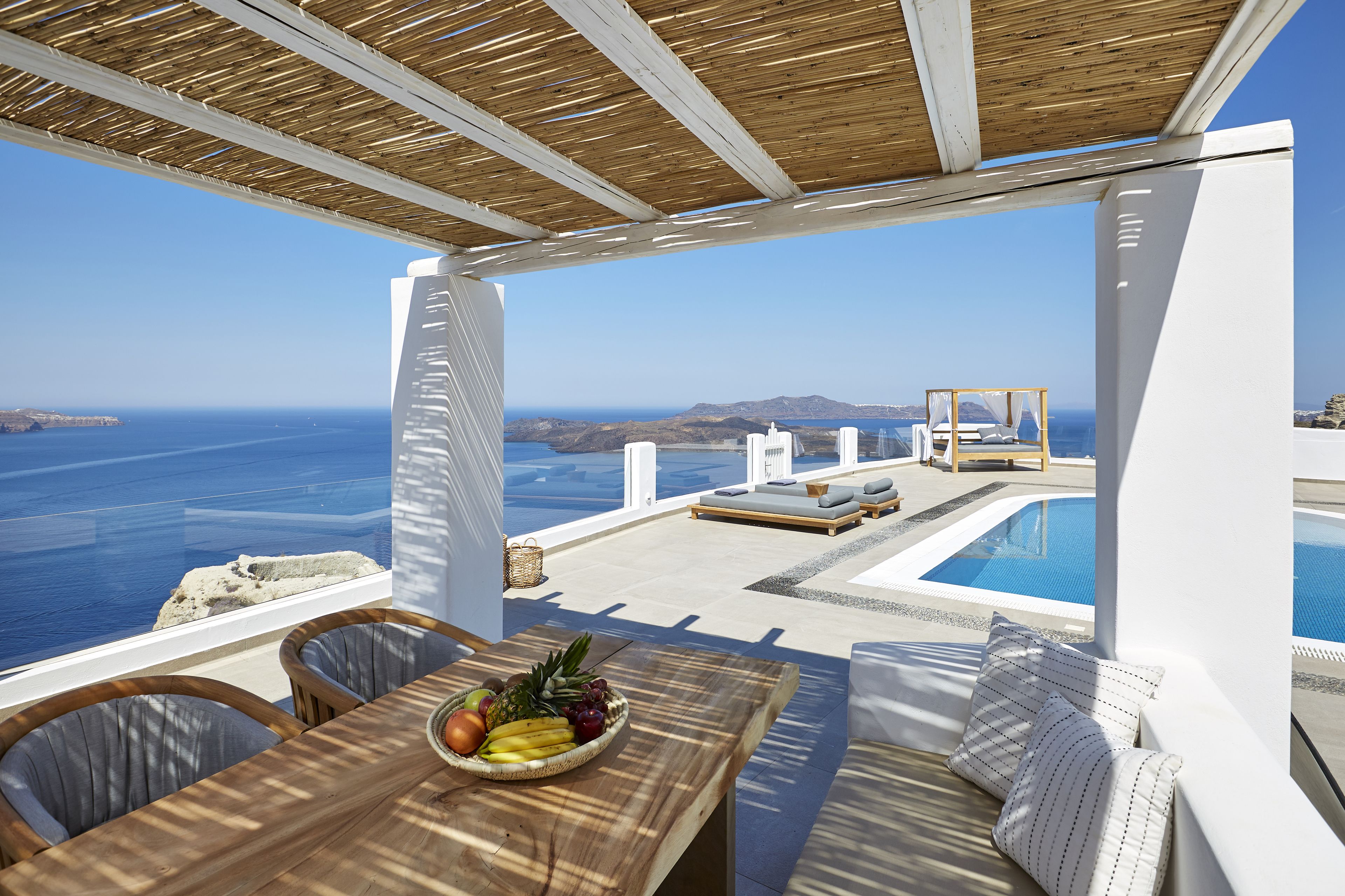 A terrace with a wooden table, a bowl of fruit, and a pool area with lounge chairs and a view of the sea.