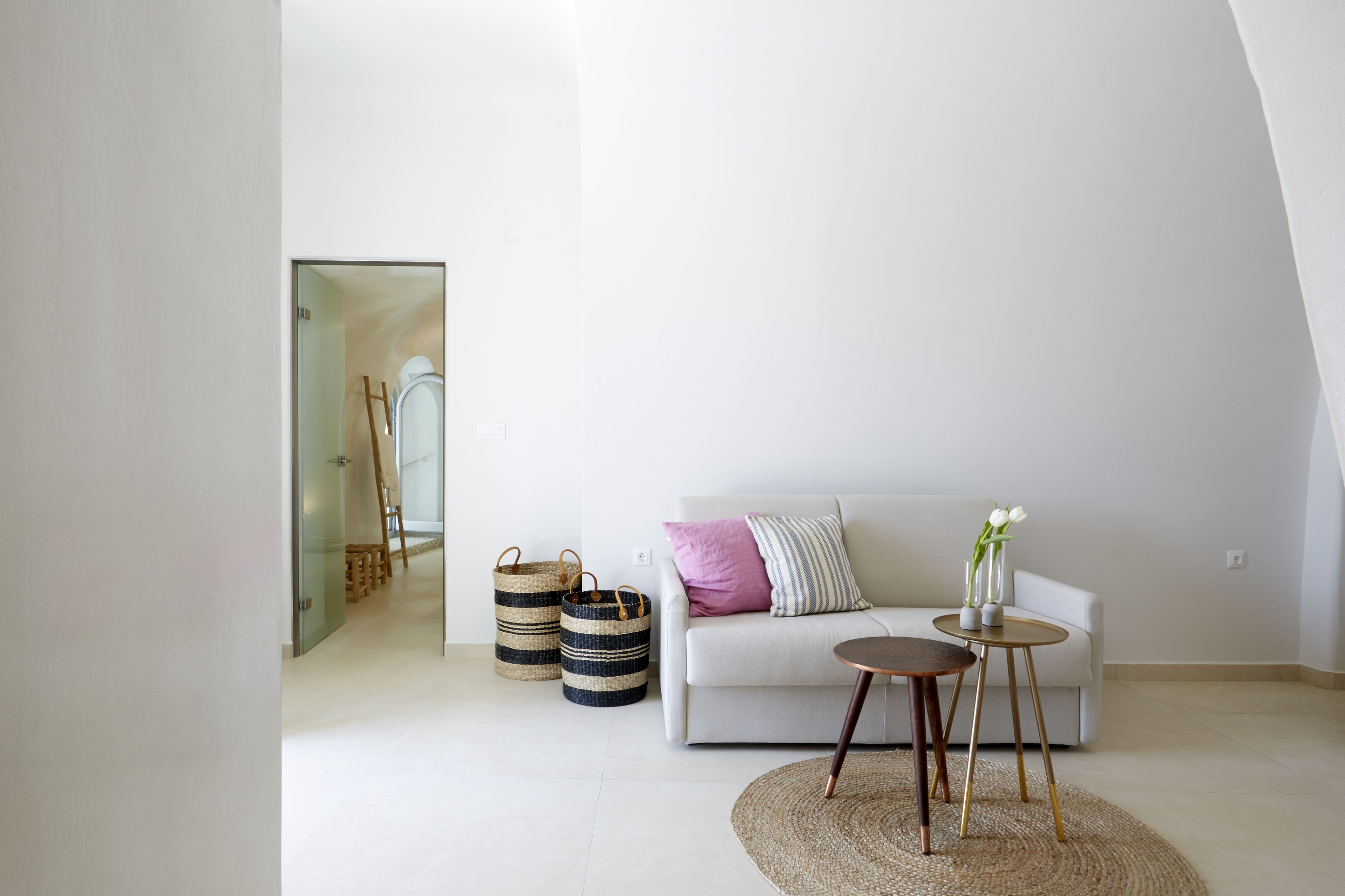 A modern living room with a white sofa, a round coffee table, and a glass door leading to another room. There are two woven baskets and a striped pillow on the sofa.
