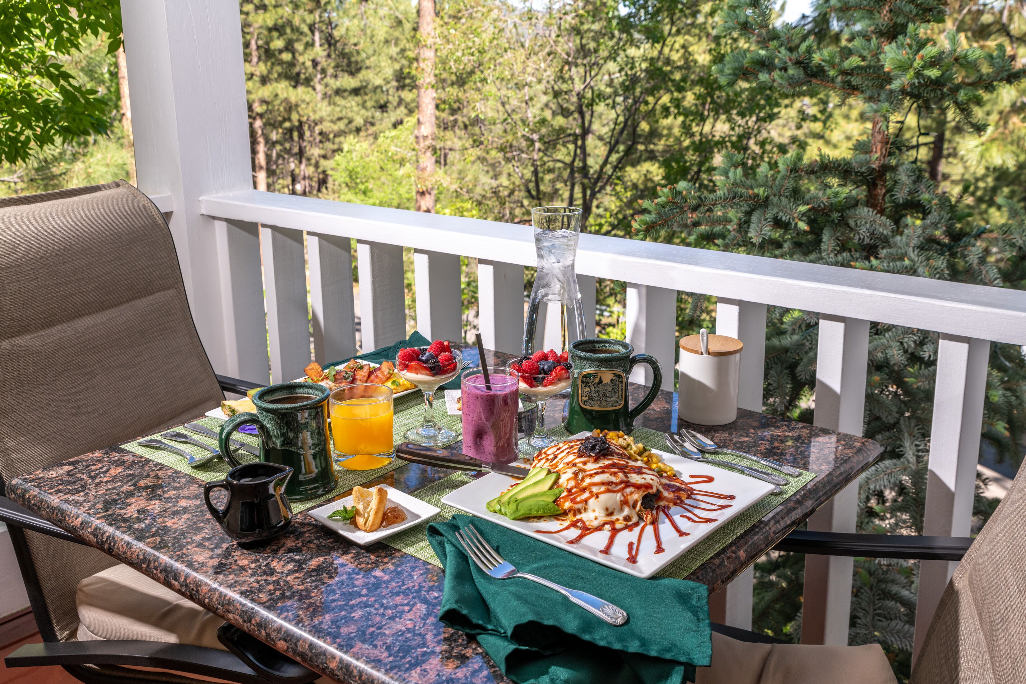 A breakfast table with a variety of foods and drinks on a balcony with a white railing and a view of trees.