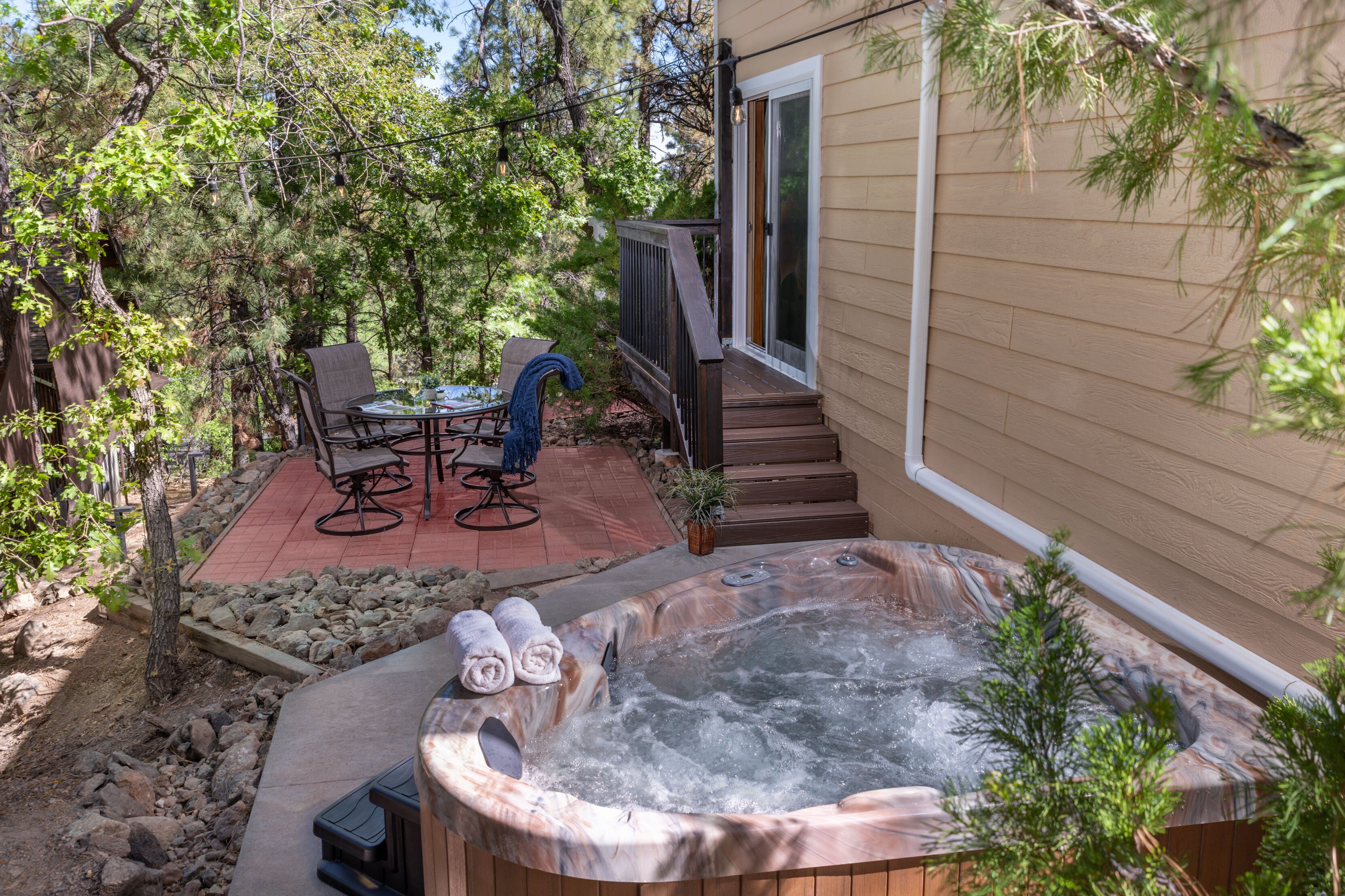 A hot tub in a backyard with a patio table and chairs, surrounded by trees and a wooden deck.
