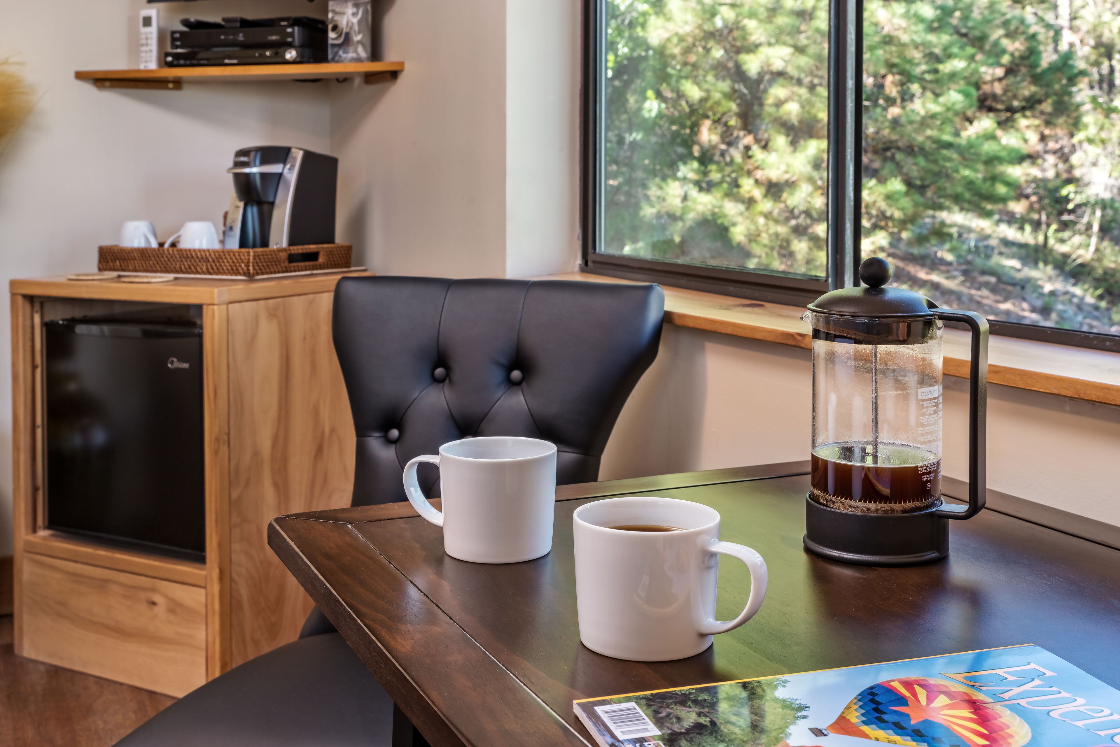 A wooden table with two white mugs, a coffee maker, and a magazine.