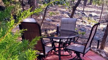 A patio with a round glass table and four chairs, surrounded by greenery and trees.