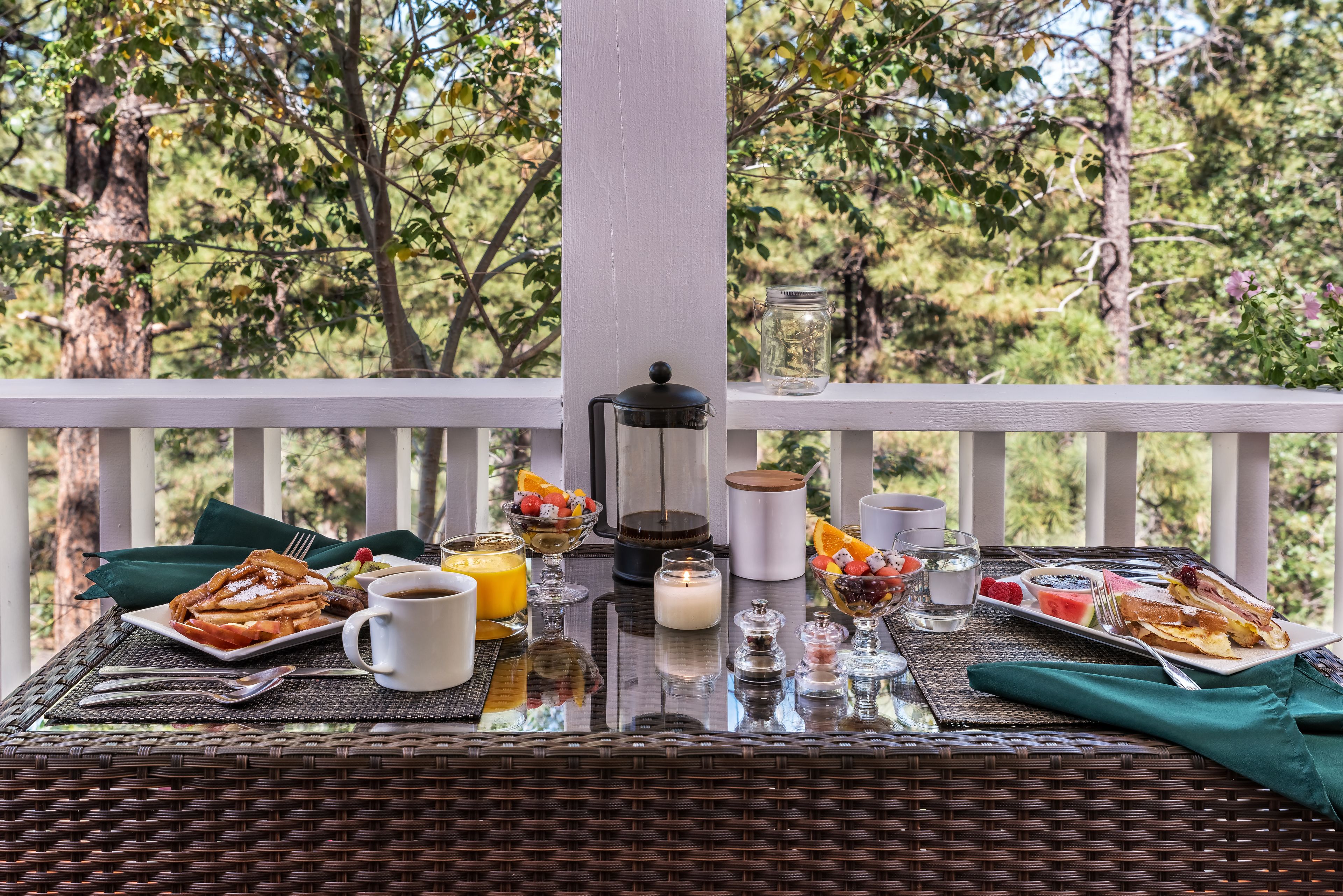 Un desayuno preparado sobre una mesa de mimbre con una barandilla blanca, que incluye una porción de tostadas francesas, un bol de fruta, una taza de café y una vela.