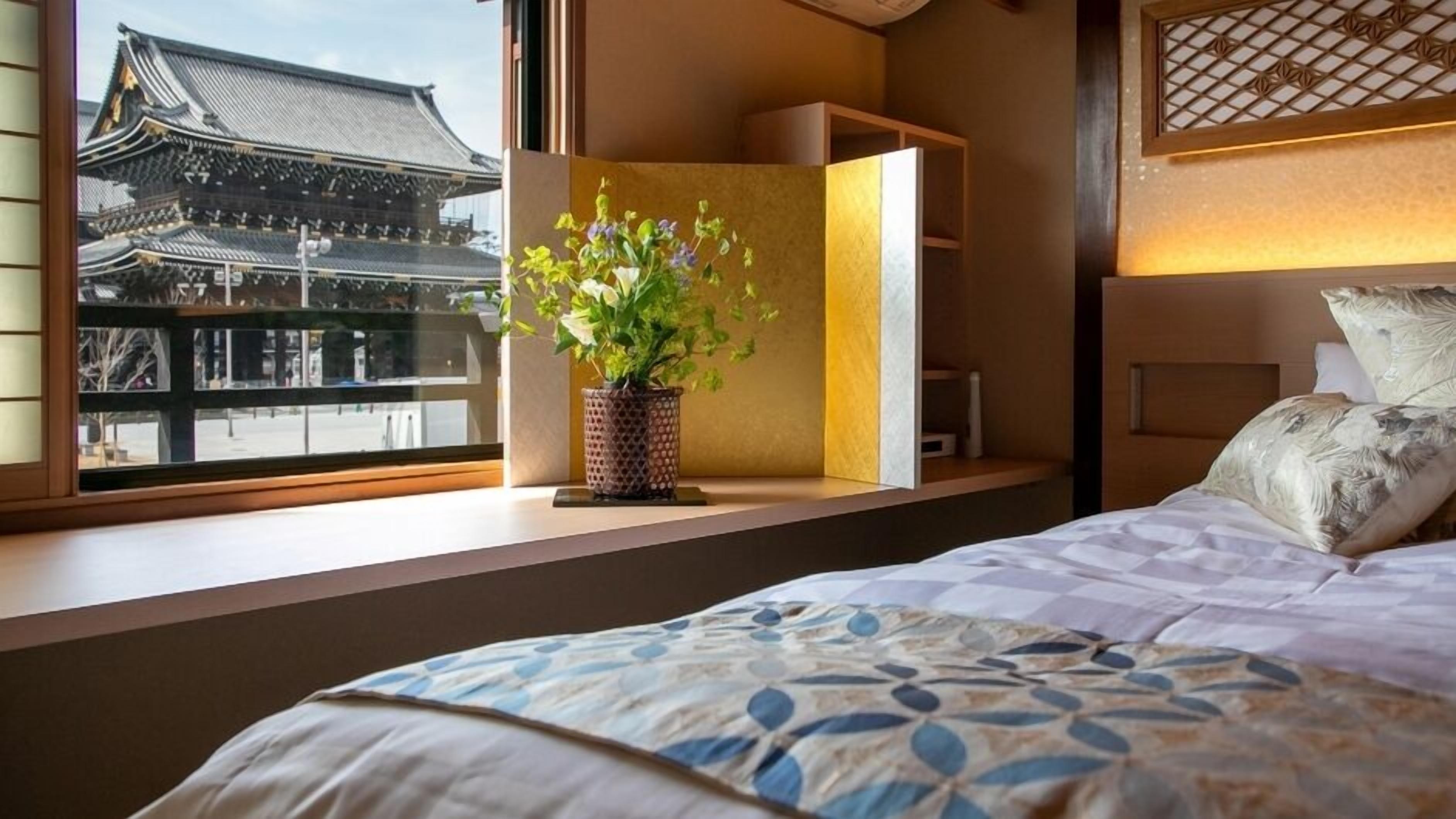 A hotel room with a bed, a window with a view of traditional architecture, a bedside table with a plant, and a headboard with a decorative panel.
