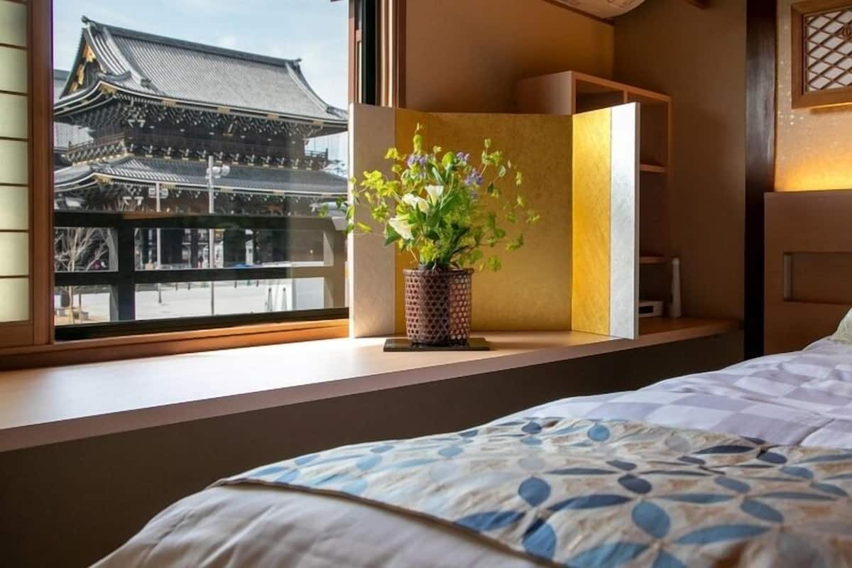 Quarto de hotel com cama, janela com vista para arquitetura tradicional, mesa de cabeceira com planta e cabeceira com painel decorativo.