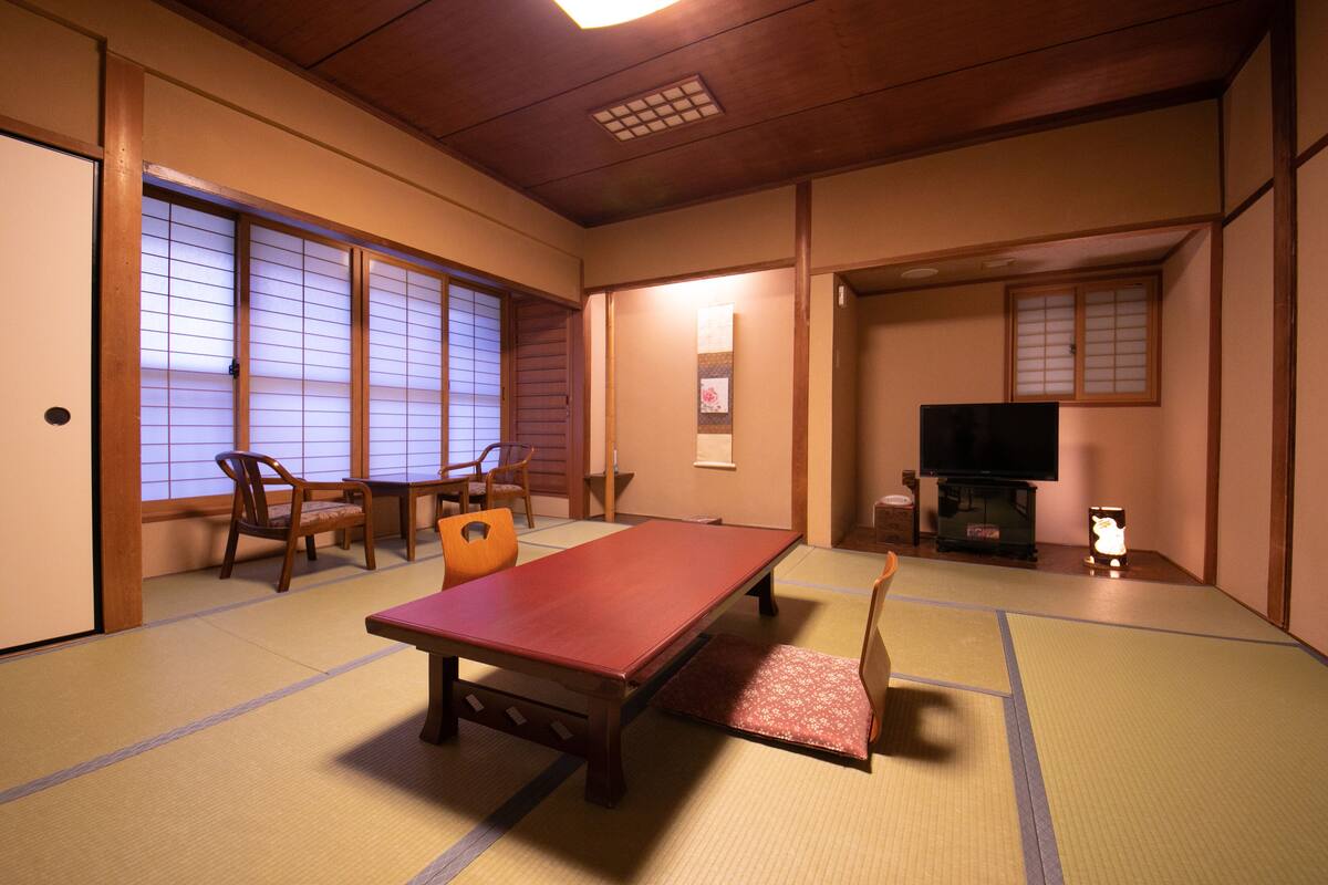 Una habitación japonesa tradicional con suelo de tatami, una mesa baja, sillas, un televisor y puertas correderas.