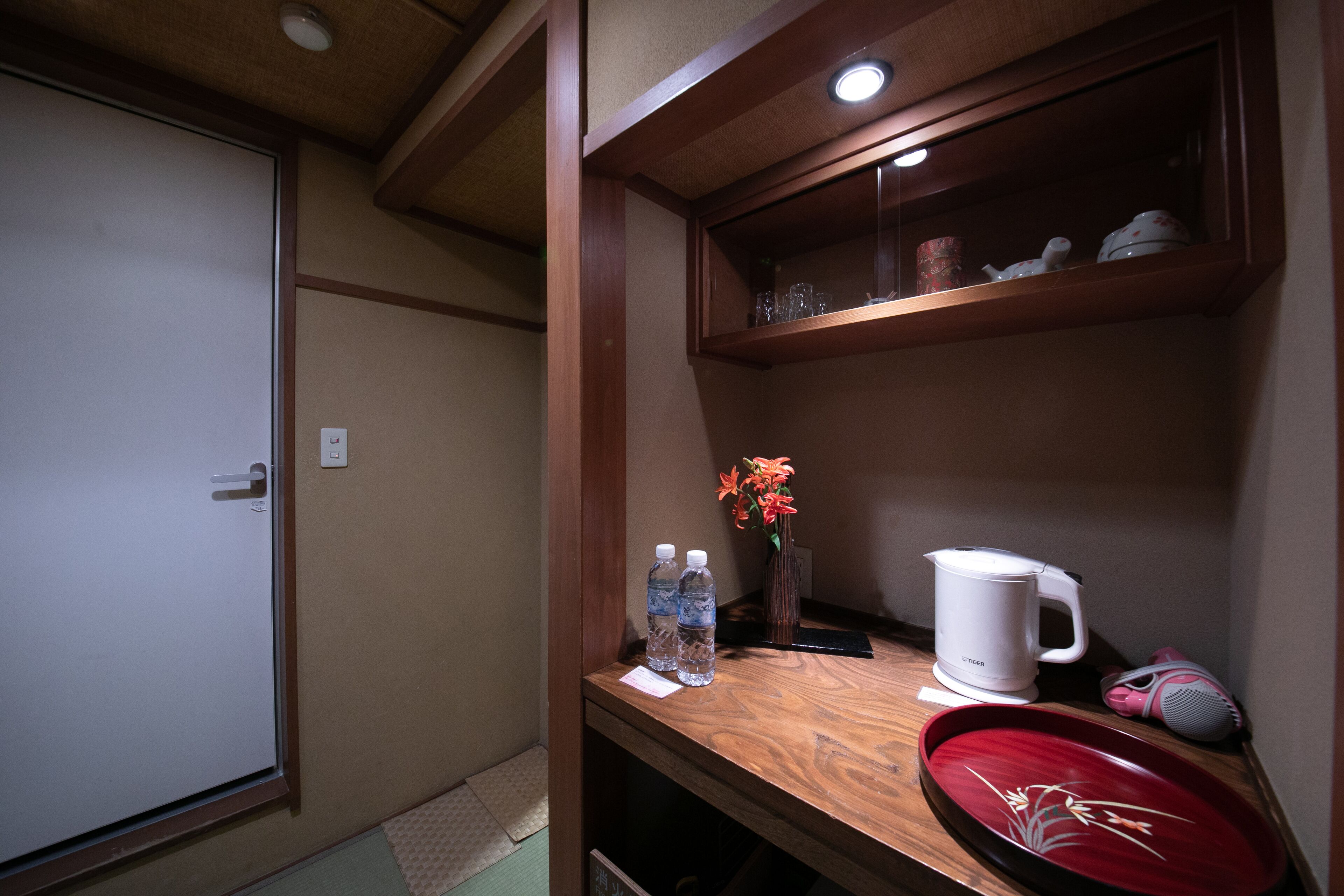 A compact room with a wooden shelf containing a kettle, a vase with flowers, and a cup. There are two water bottles on the shelf, and a small tray with a floral design.