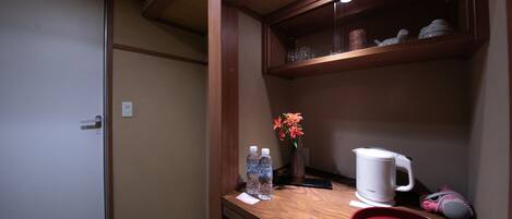 A compact room with a wooden shelf containing a kettle, a vase with flowers, and a cup. There are two water bottles on the shelf, and a small tray with a floral design.