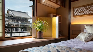 A hotel room with a large window offering a view of traditional architecture, a bedside table with a potted plant, and a bed with a patterned bedspread. - Hokkaikan Ohanabo (Kyoto)
