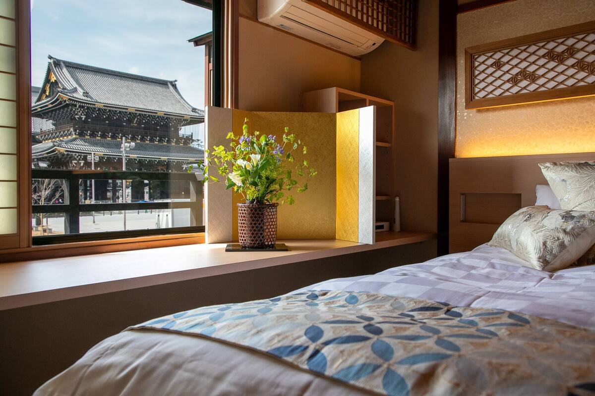 Habitación de hotel con un ventanal grande que ofrece vistas de arquitectura tradicional, una mesita de noche con planta en maceta y una cama con edredón estampado.