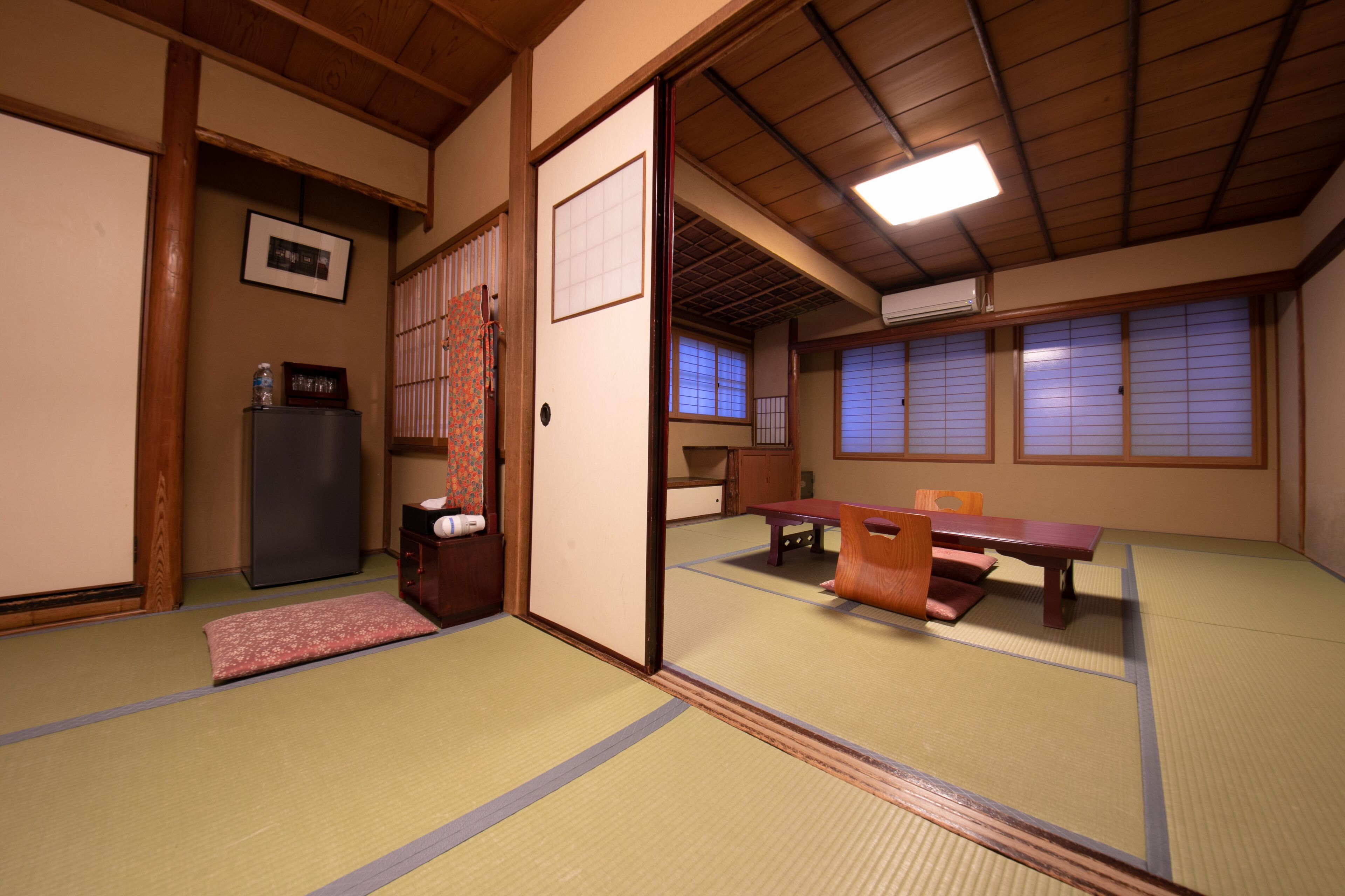 A traditional Japanese room with tatami flooring, a low table with cushions, a sliding door, and a refrigerator.
