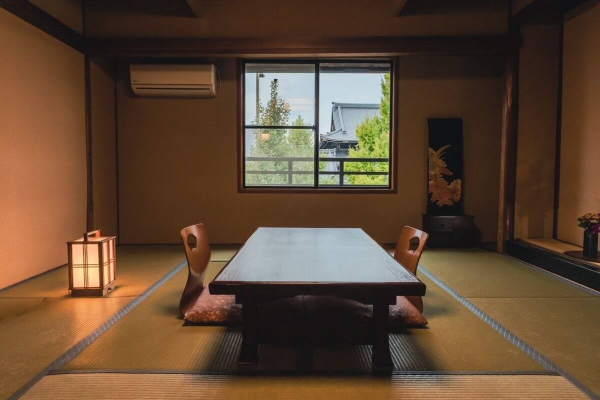 Una habitación japonesa tradicional con suelo de tatami, una mesa baja y una ventana que ofrece vistas a un edificio tradicional.