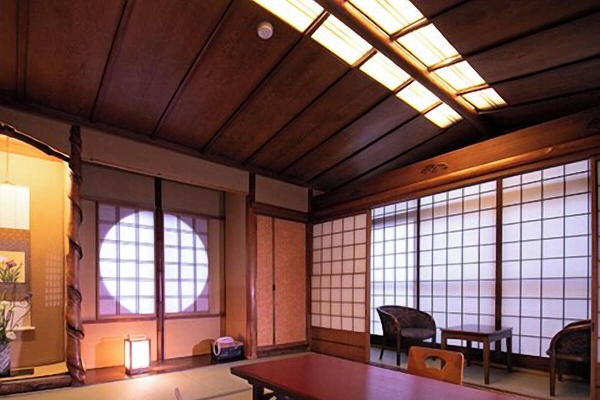 Una habitación con esterillas de tatami, una mesa baja y sillas, una puerta corredera y vistas a través de un mampara de shoji.