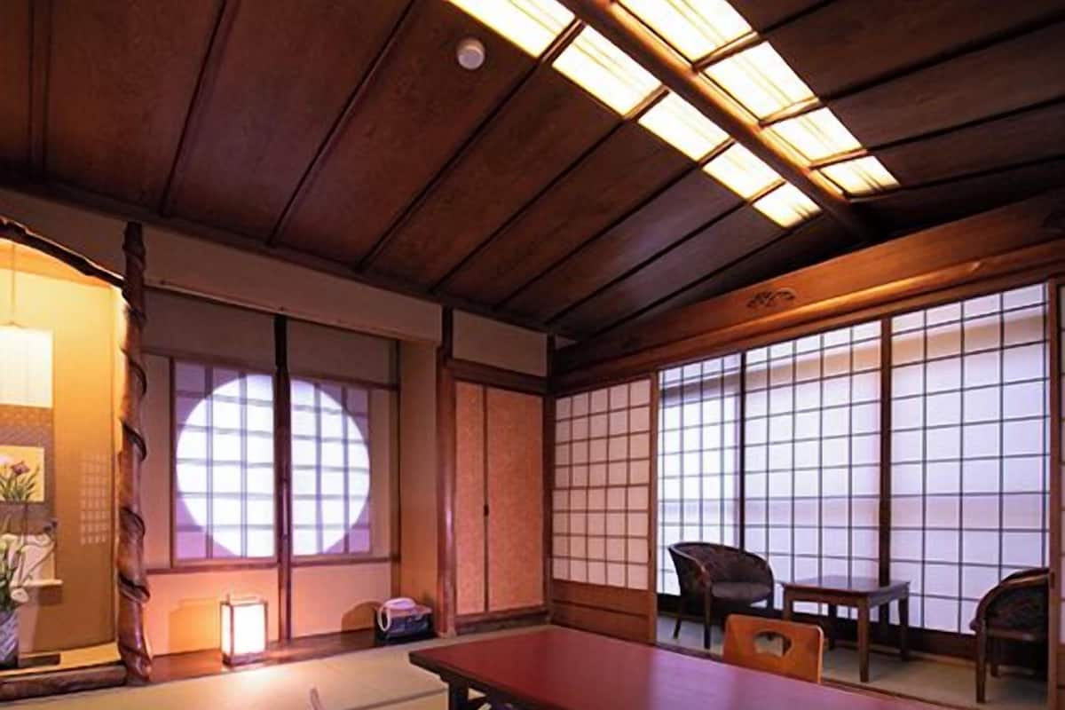 Ein Raum mit Tatami-Matten, einem niedrigen Tisch, Stühlen, einer Schiebetür und Blick durch einen Shoji-Bildschirm.