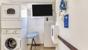 A laundry room with a washing machine, dryer, ironing board, and a television mounted on the wall.