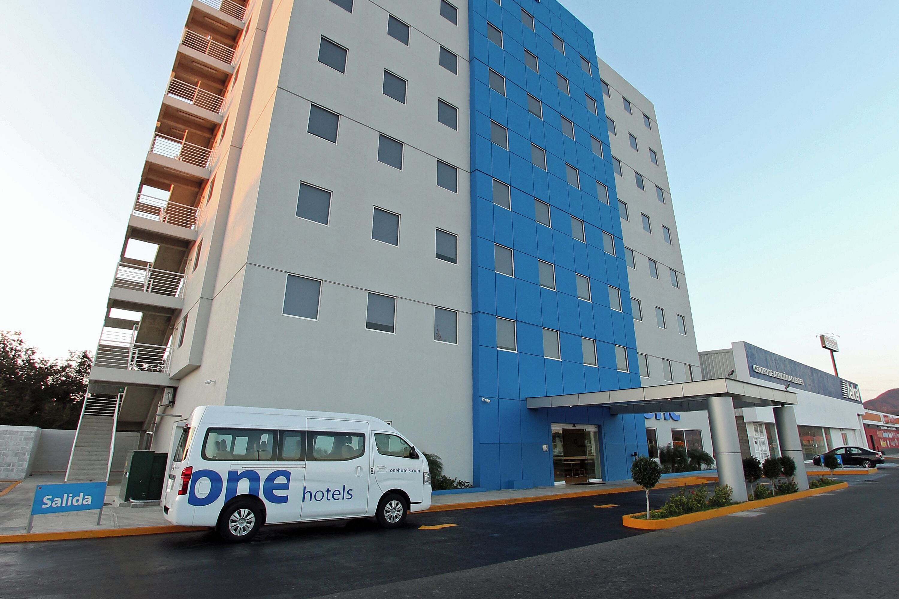 Un hôtel moderne, avec une façade blanche et bleue, une entrée couverte et un fourgon garé devant.