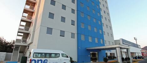 A modern hotel building with a white and blue facade, a covered entrance, and a van parked in front.
