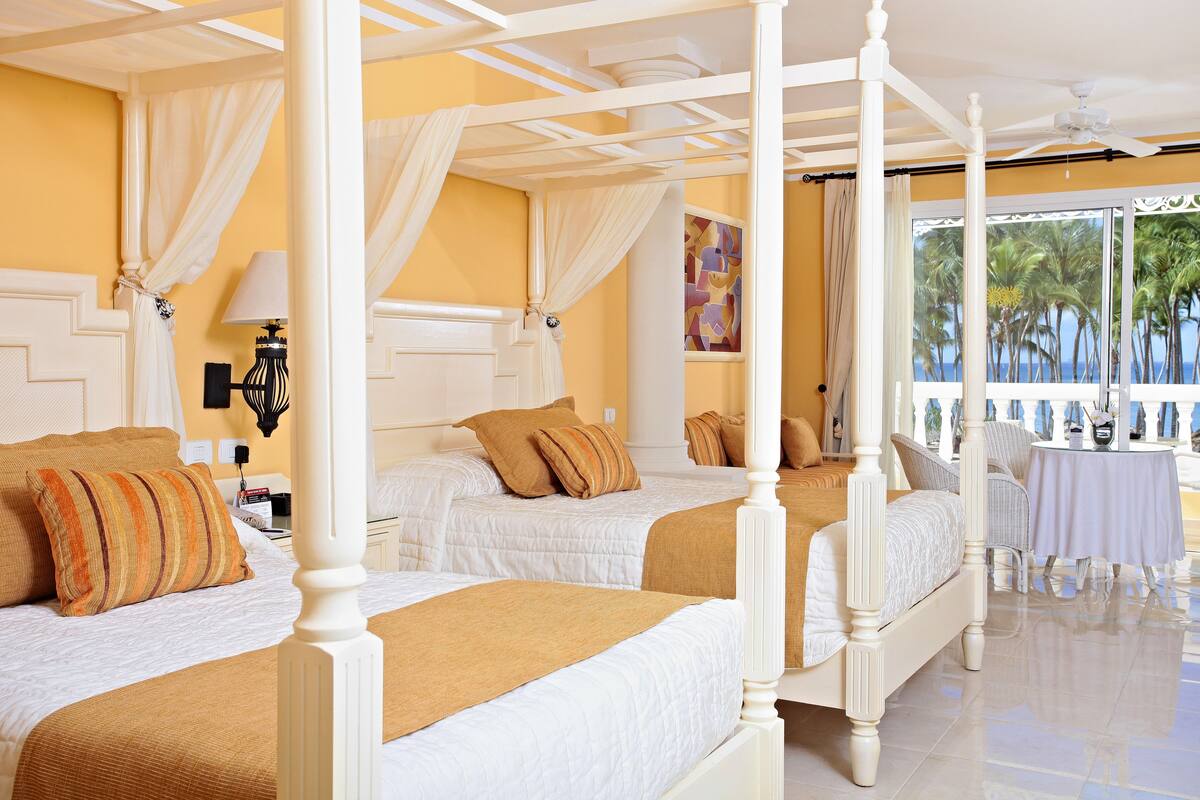 Una cama con dosel, ropa de cama blanca y un plaid color beige. En primer plano, hay una pequeña mesa con mantel blanco y una silla. La habitación tiene una pared amarilla y un ventanal con vistas a palmeras.