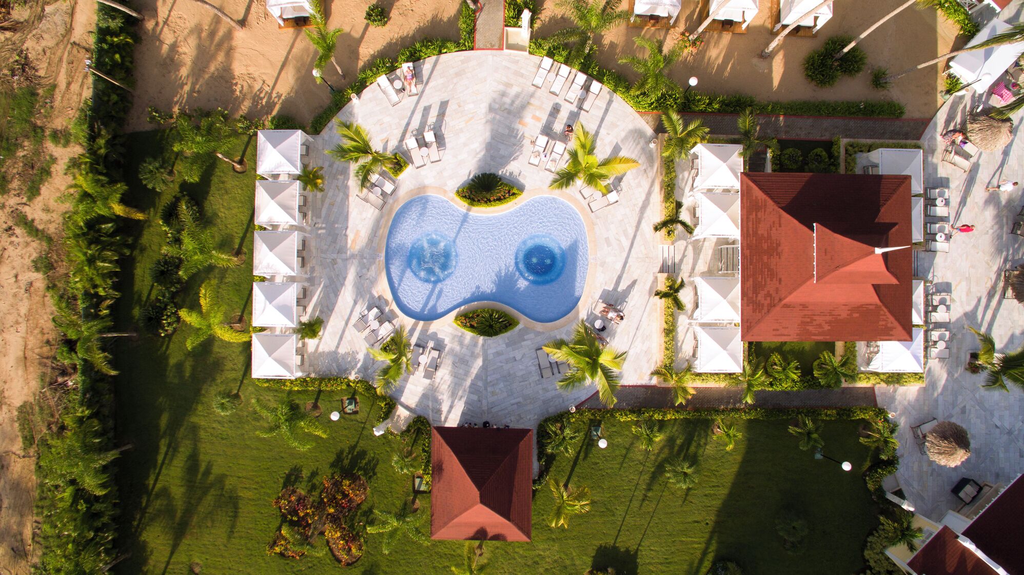 Vista aérea de un complejo turístico con una piscina central, rodeado de zonas de descanso y palmeras.