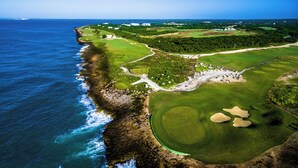 An aerial view of a coastal golf course with a large water body to the left, a well-maintained green, and a few sand bunkers.