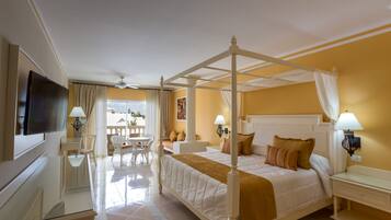 A four-poster bed with white bedding, a television, a desk with a lamp, and a chair in a room with yellow walls and a balcony view.