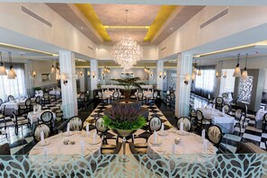 Une salle à manger élégante, dotée d’un lustre central, de plusieurs tables et de chaises disposées pour le service.