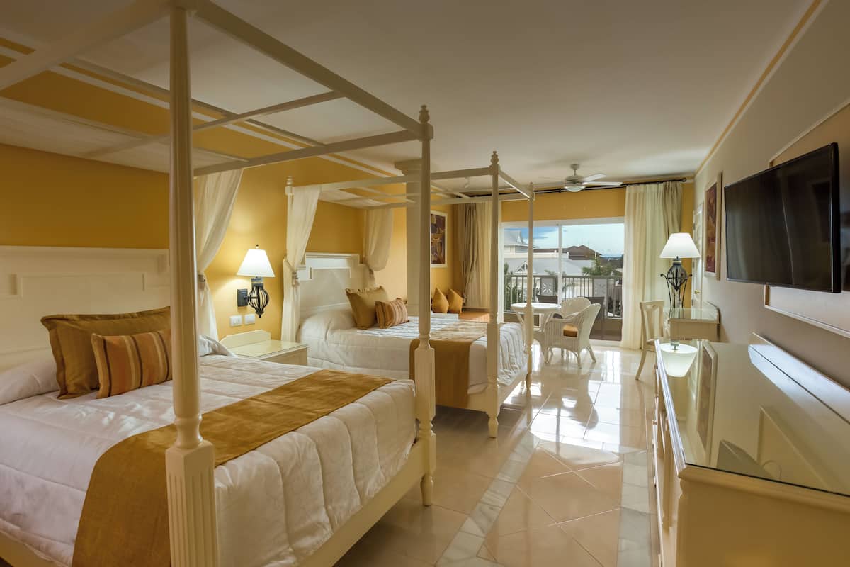 Habitación de hotel con cuatro camas dispuestas en forma de U, un televisor en la pared y un balcón con vistas.