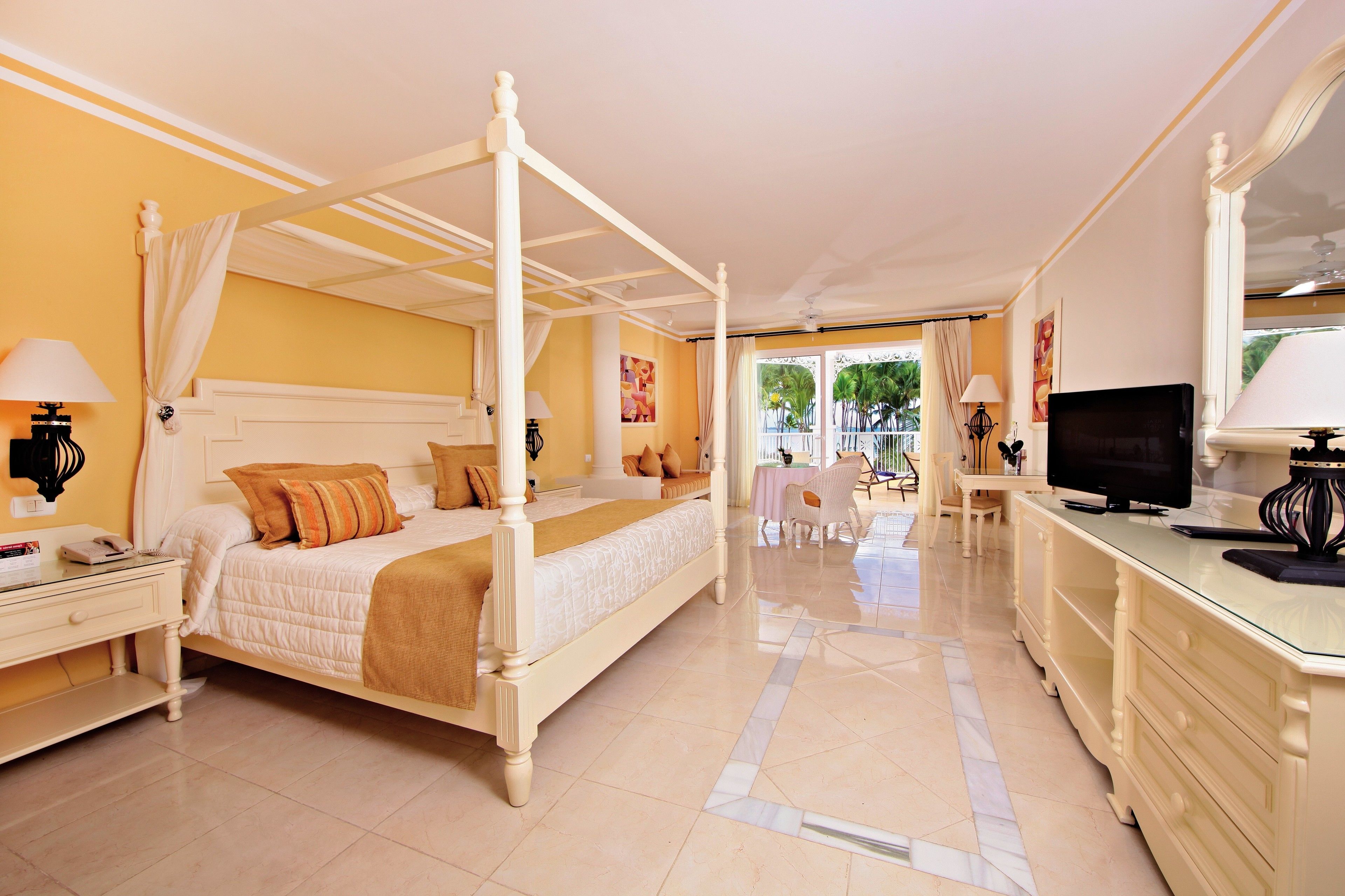 A four-poster bed with a beige bedspread and pillows, a television on a glass-topped table, and a desk with a lamp.