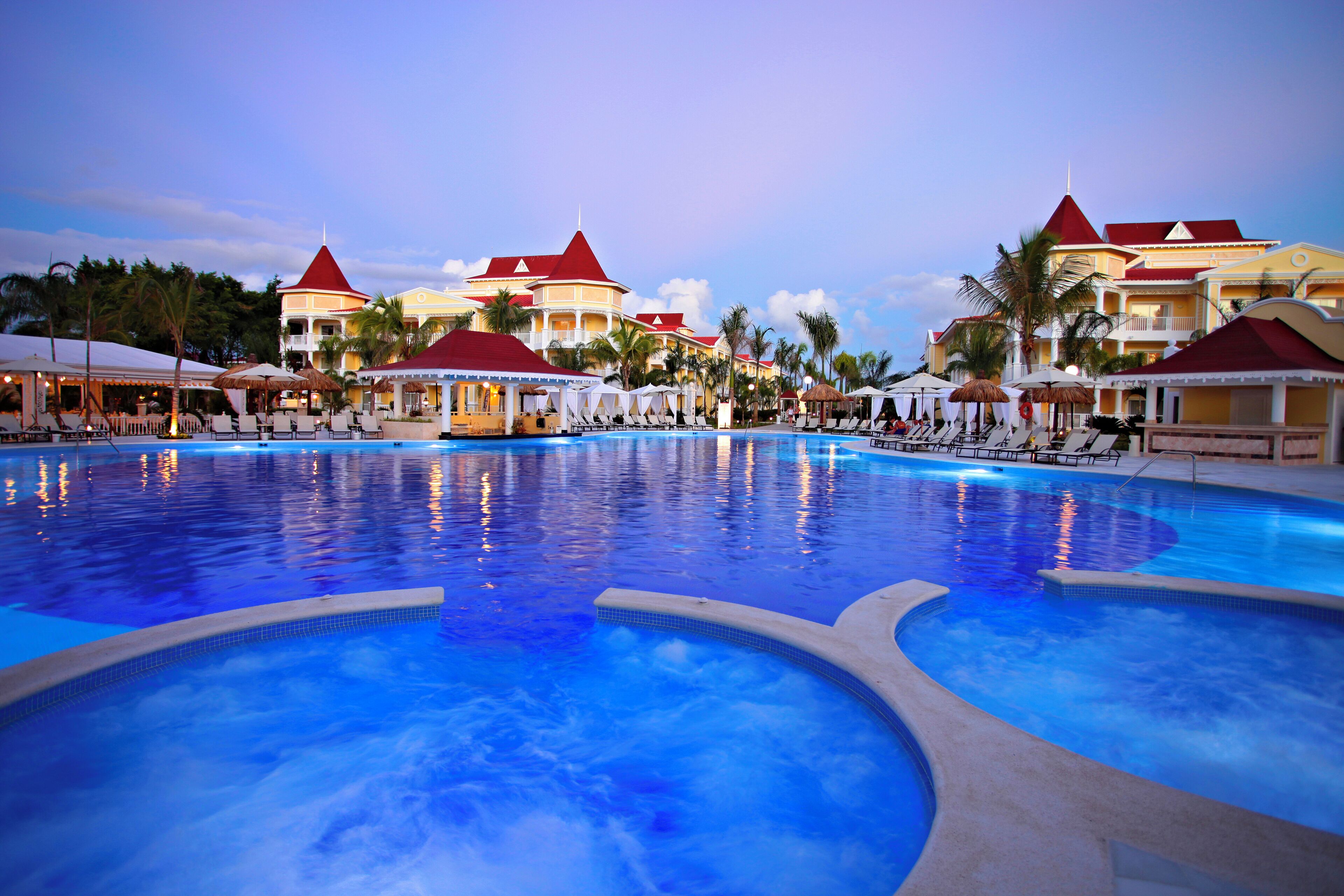A large swimming pool with lounge chairs and umbrellas, surrounded by resort buildings with red roofs.
