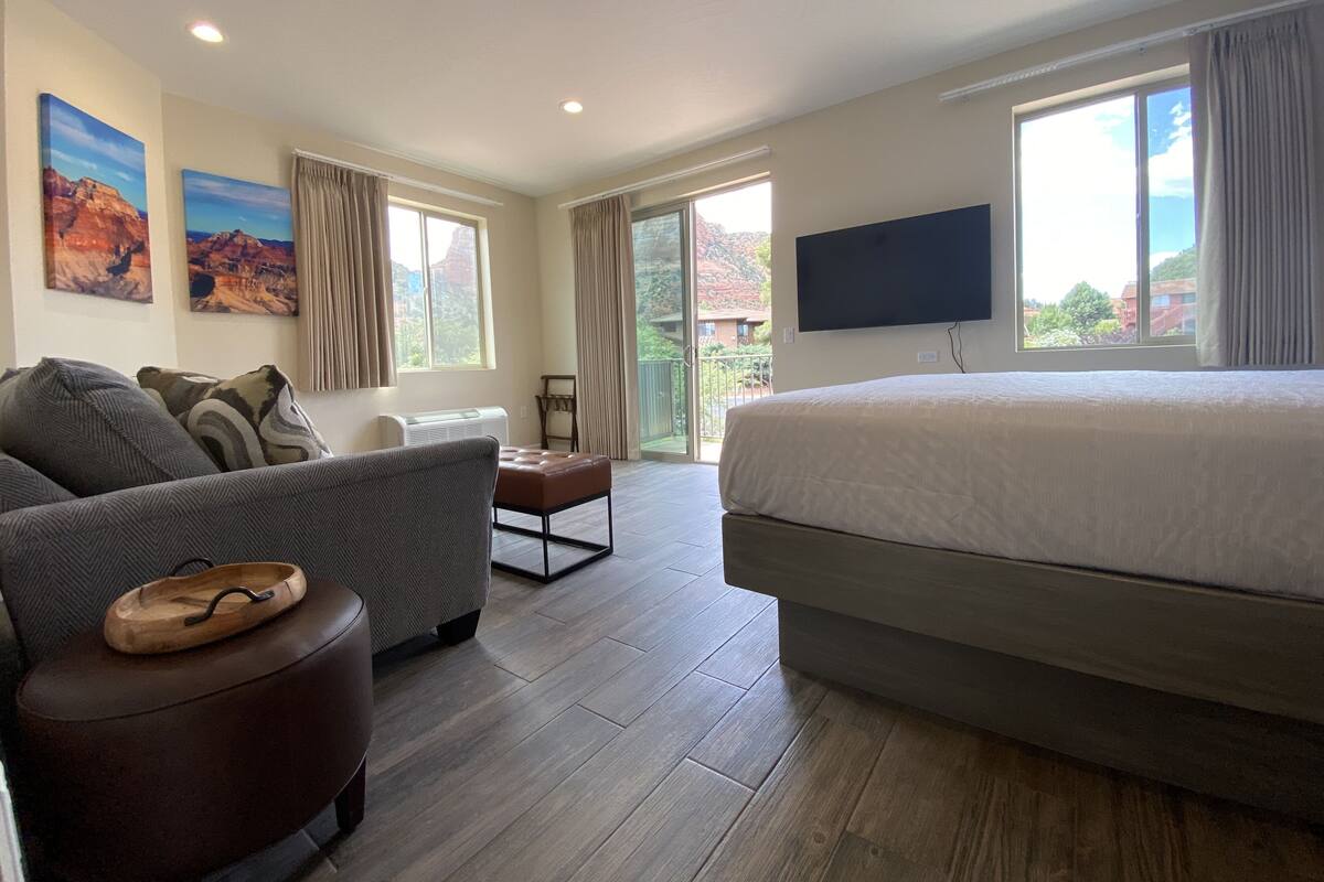 Ein Hotelzimmer mit einem großen Bett, einer Couch, einem kleinen Tisch und einem Fernseher. Zwei Fenster mit Vorhängen, und ein Balkon ist durch die Fenster sichtbar.