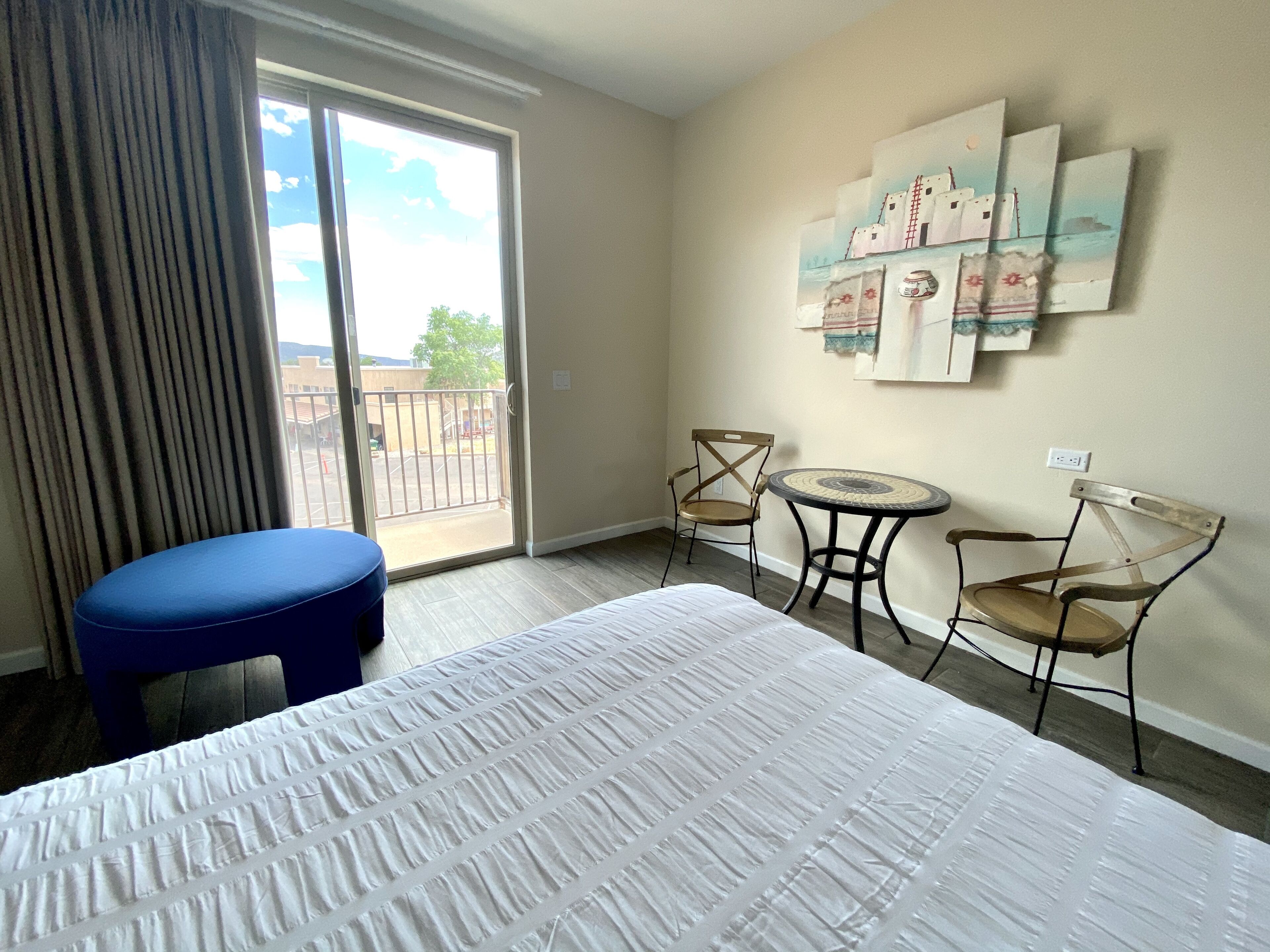 Habitación de hotel con cama, un taburete azul, dos sillas, una mesita redonda y un balcón con vistas al exterior.
