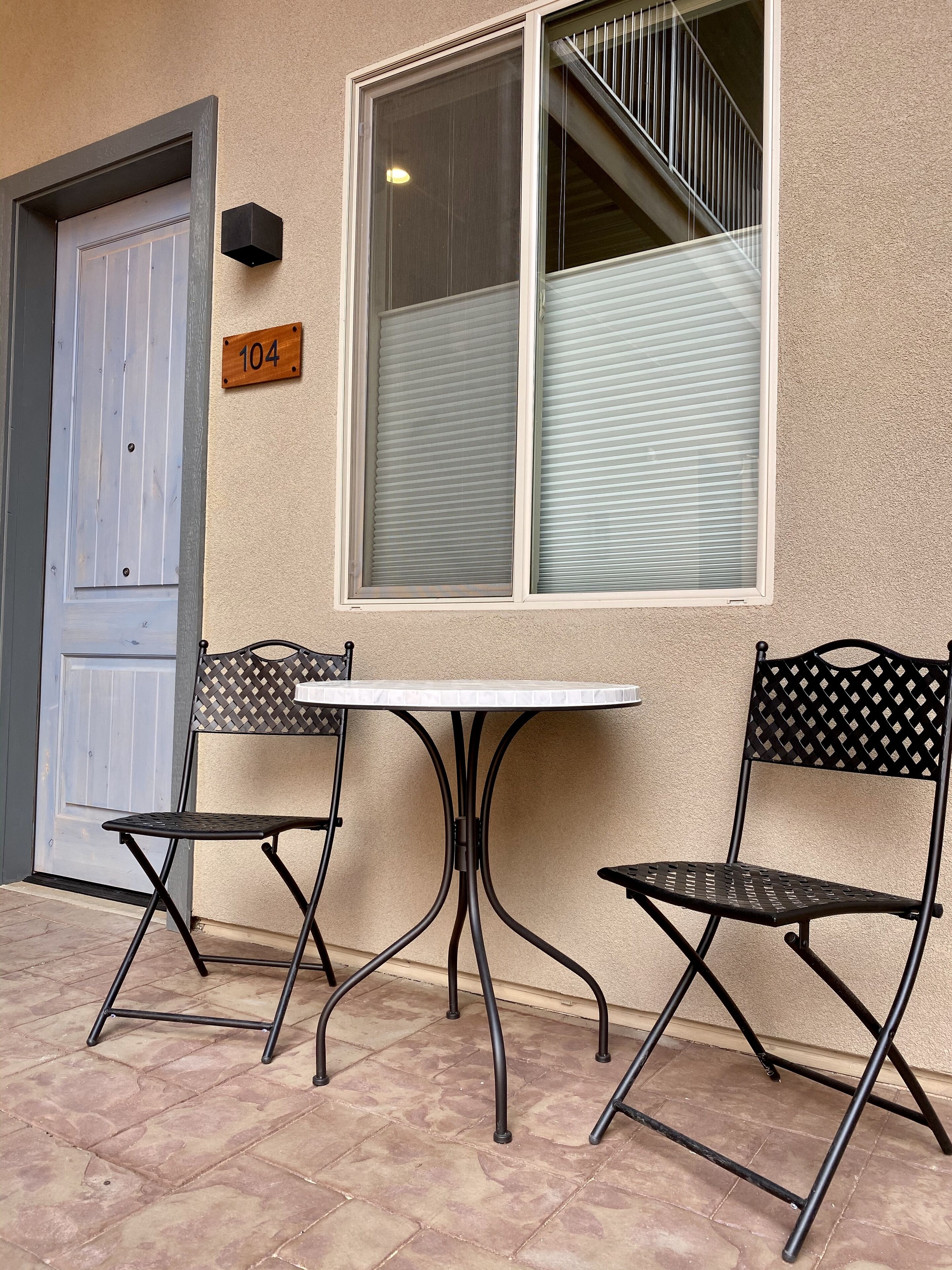 A building exterior with a door, a window with blinds, and two metal chairs with a table in front.