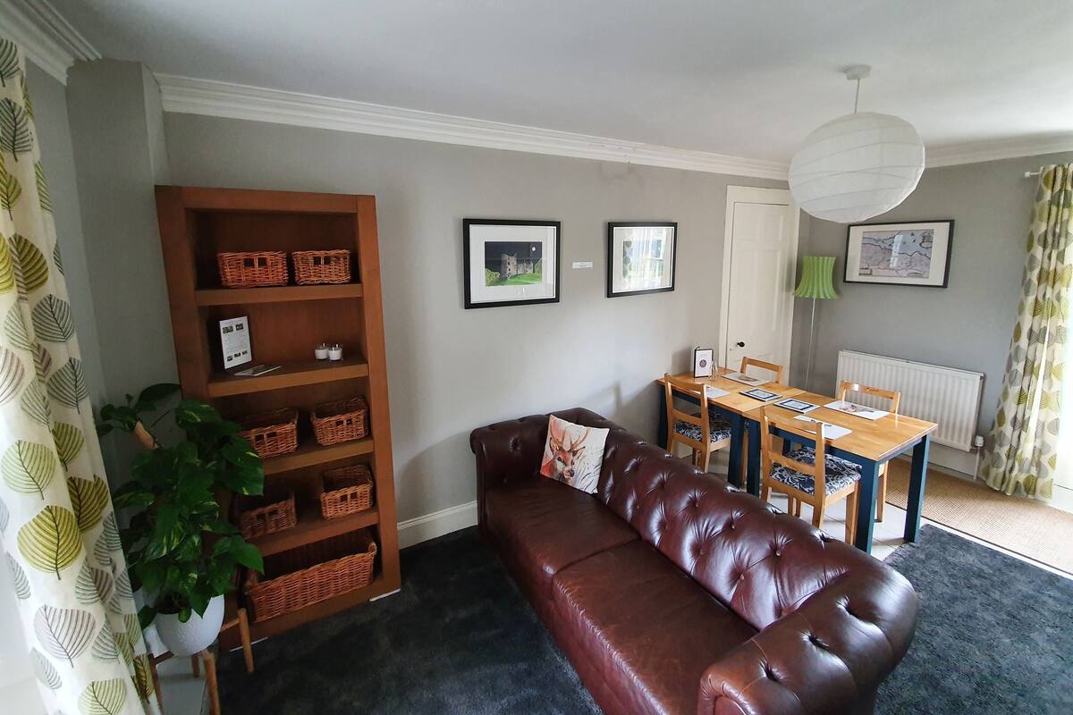 Een woonkamer met een bruine leren bank, een houten boekenkast, een eettafel met stoelen, en ingelijste foto's aan de muur.