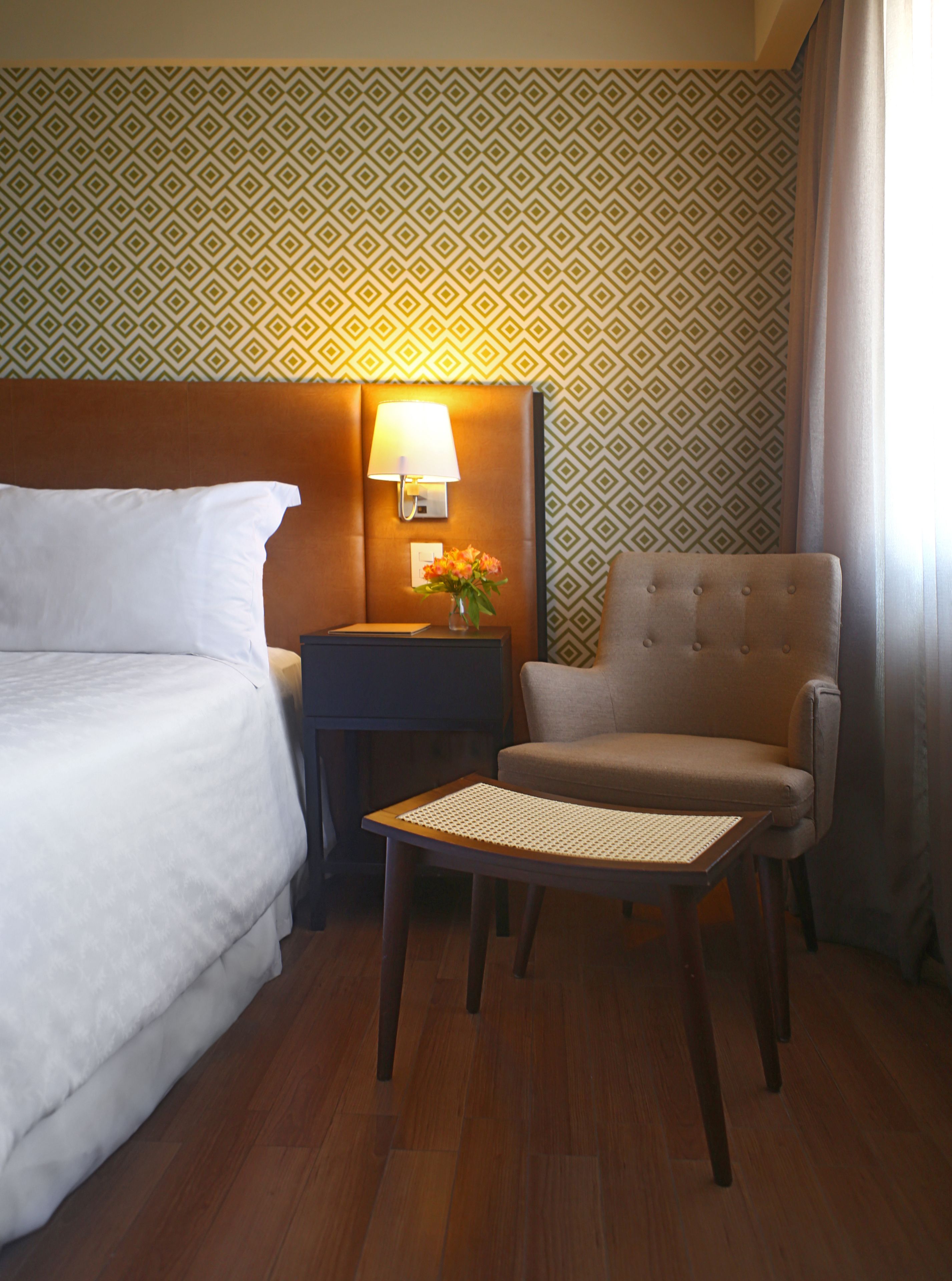 Una camera d'hotel con un letto, una poltrona, un tavolino, un comodino con una lampada e carta da parati decorata.