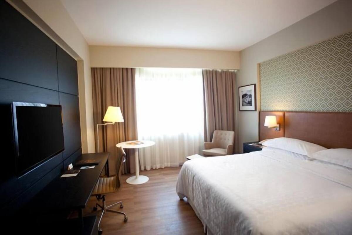 Une chambre d’hôtel équipée d’un lit, d’une télévision, d’un bureau, d’une chaise, d’une lampe et d’une fenêtre avec des rideaux.