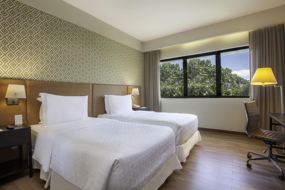 Une chambre d’hôtel avec deux lits, un bureau, une chaise et une fenêtre donnant sur des arbres.