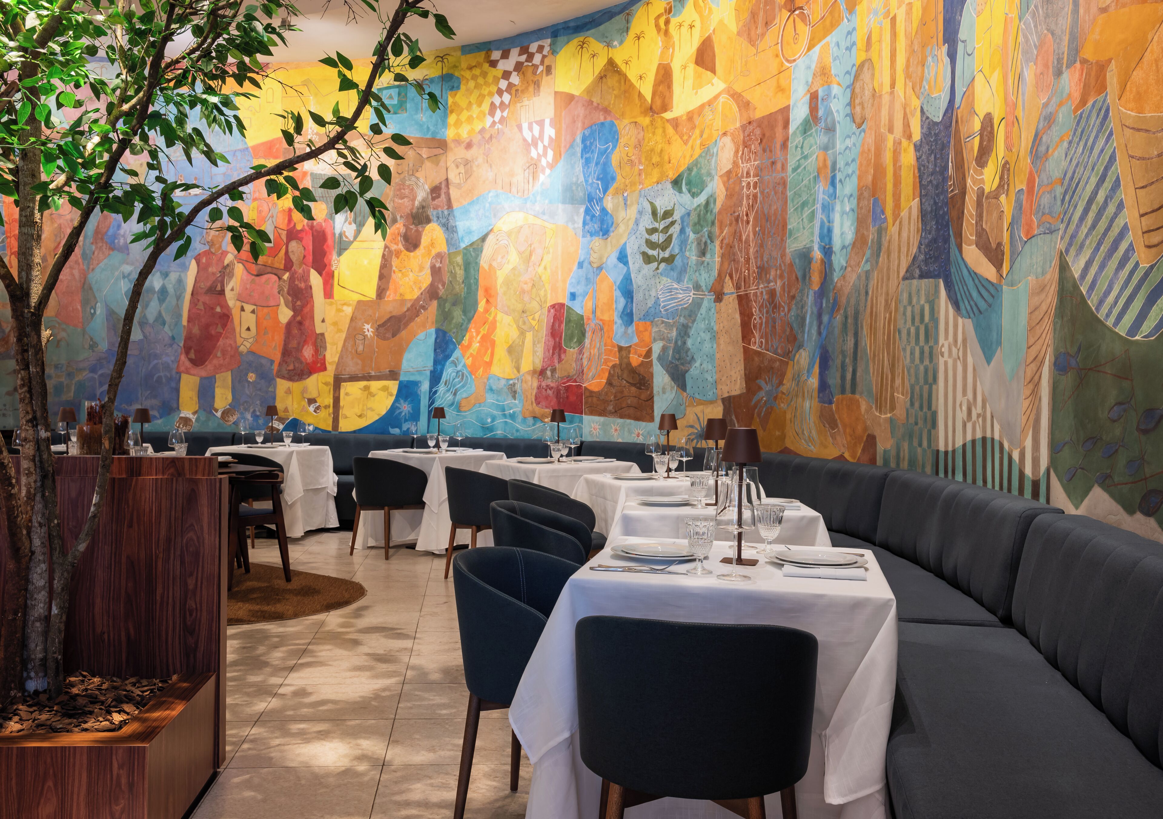 Un restaurante con mesas y sillas, un mural en la pared y un árbol en la esquina.